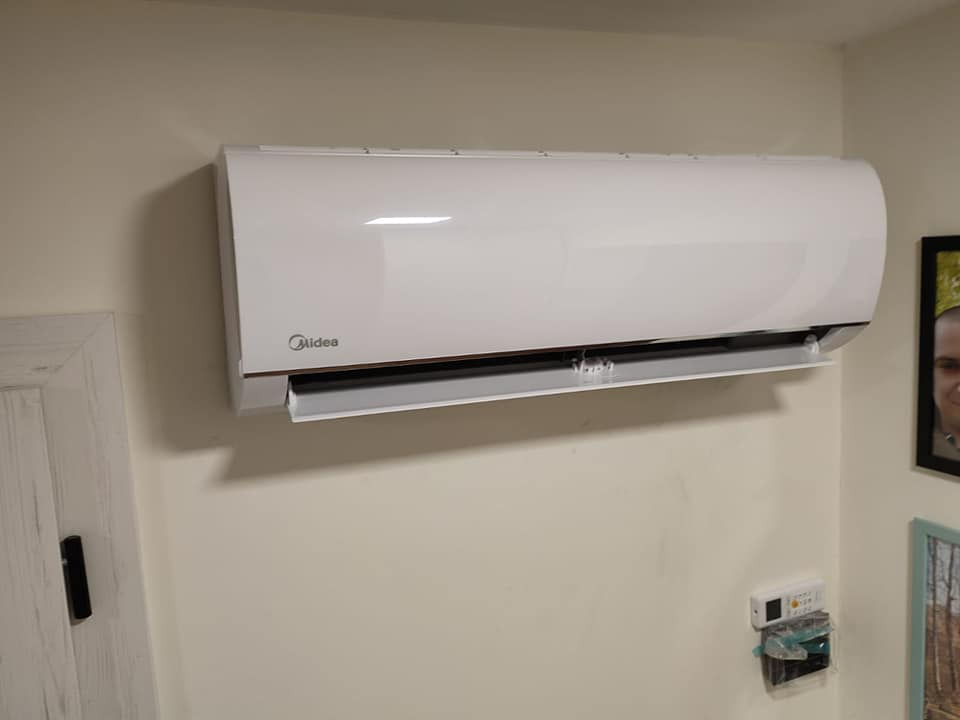 Biały klimatyzator Midea zamontowany na jasnej ścianie w pomieszczeniu, widoczny pilot na ścianie i fragment zdjęcia w ramce.
