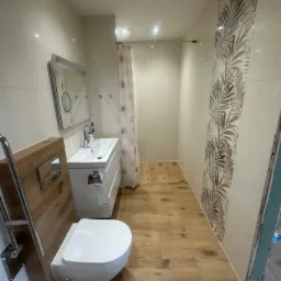 Wykończona łazienka z drewnopodobną podłogą, białymi płytkami i akcentem dekoracyjnym w postaci pionowego pasa płytek z motywem liści palmowych. Widoczna podwieszana toaleta, umywalka z szafką...