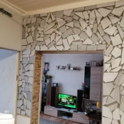 Wykończenie wnętrza z dekoracyjną ścianą z nieregularnych, jasnych płytek kamiennych, widok na pokój z meblami i telewizorem.