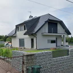 Nowoczesny dom z szarym dachem i elewacją w odcieniach bieli, otoczony gabionowym ogrodzeniem z elementami metalowymi, widok z zewnątrz w pochmurny dzień.
