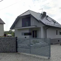 Dwukondygnacyjny dom jednorodzinny z szarą elewacją, ciemną dachówką i balkonem z szarymi żaluzjami, widoczny od strony frontu z szarym, metalowym ogrodzeniem panelowym i gabionami.