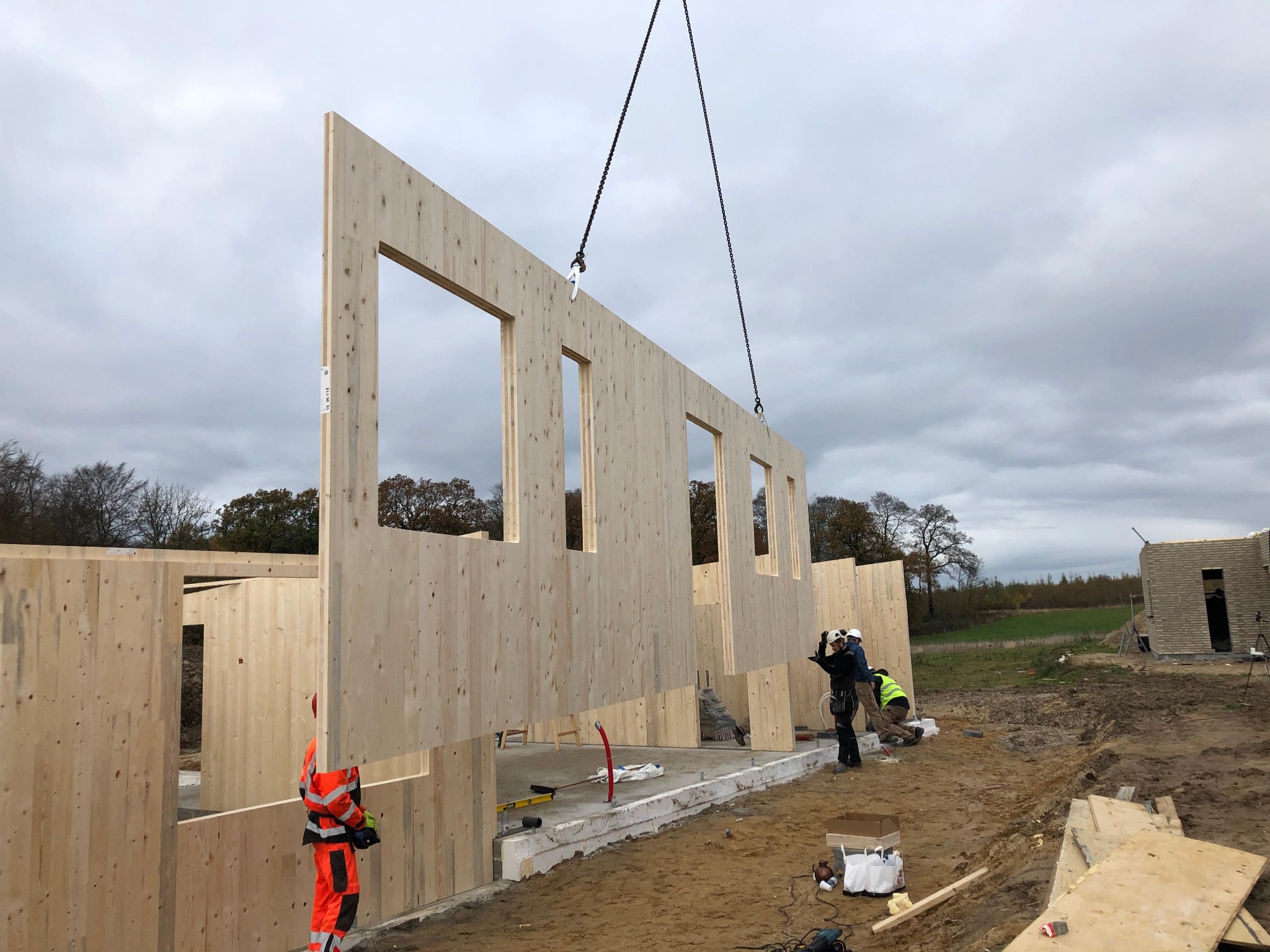 Montaż ściany z prefabrykatów drewnianych z otworami okiennymi za pomocą dźwigu na placu budowy, widoczni pracownicy w odzieży roboczej.