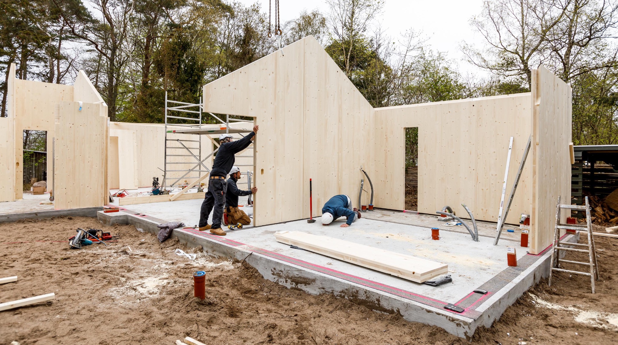 Montaż prefabrykowanych ścian domu z drewna klejonego warstwowo; widoczni trzej pracownicy w kaskach, poziomica oparta o ścianę, teren budowy w tle.