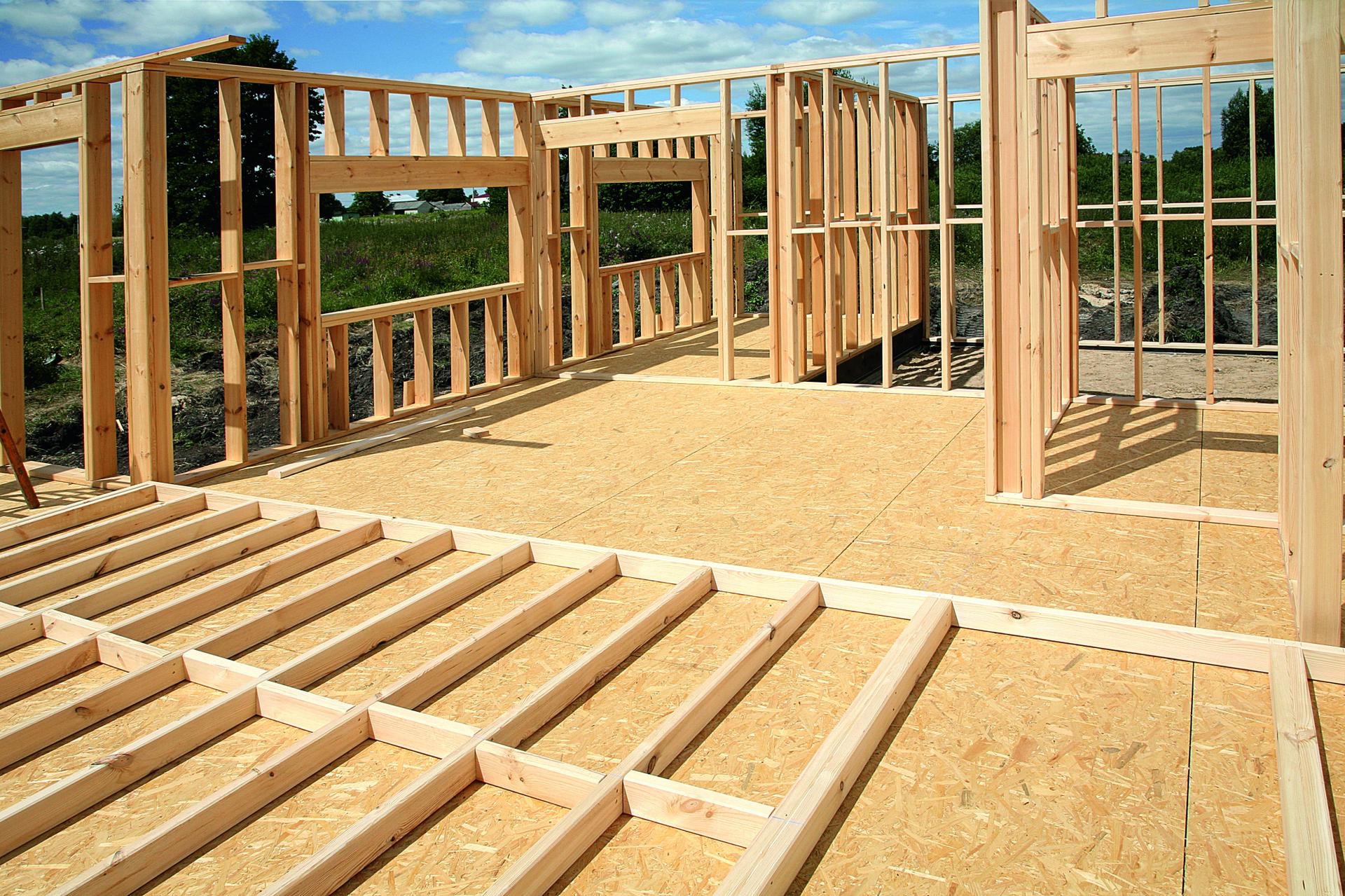 Wnętrze domu szkieletowego w trakcie budowy, widoczne drewniane ramy ścian i podłogi na płycie OSB, z zielonym krajobrazem w tle przez otwory okienne.