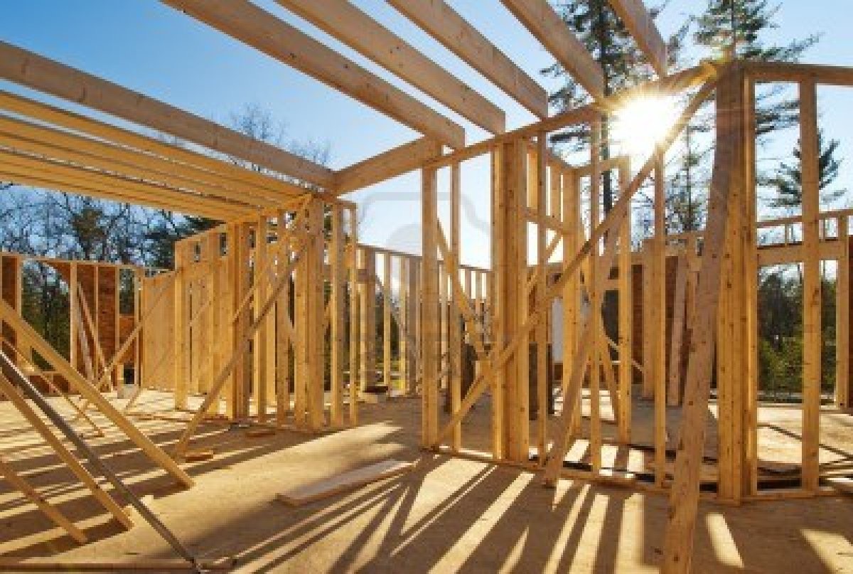 Wnętrze w trakcie budowy z drewnianą konstrukcją szkieletową w jasnym świetle słonecznym, widoczne elementy więźby dachowej i słupy nośne.