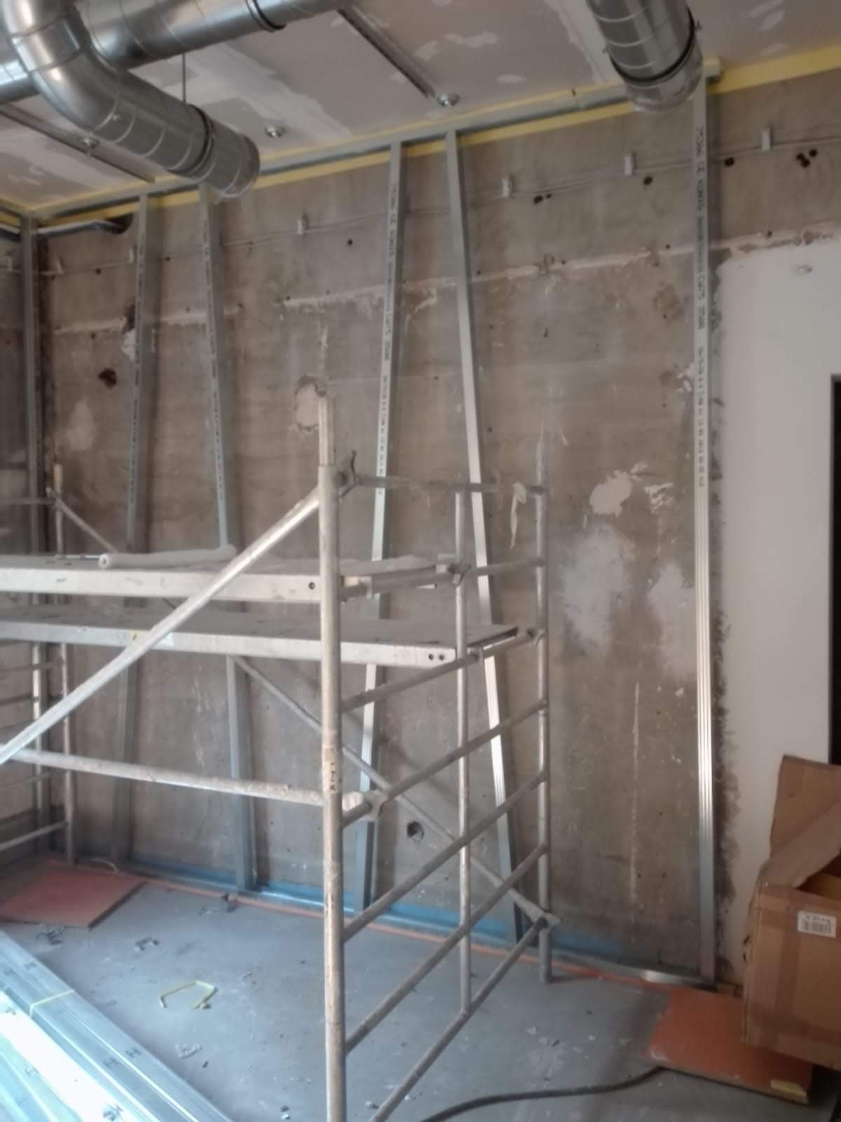 Wnętrze w trakcie prac wykończeniowych: surowa ściana z metalowymi profilami, widoczne rusztowanie i instalacja wentylacyjna pod sufitem.