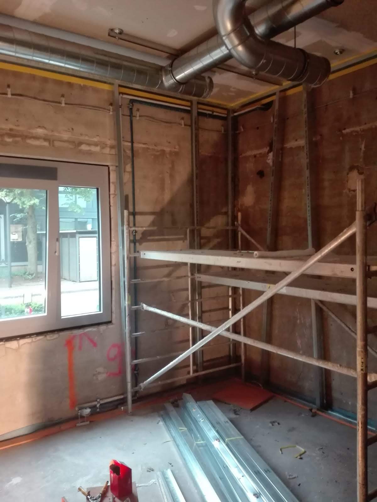 Wnętrze w trakcie remontu z widocznymi rusztowaniami, metalowymi profilami na ścianach, rurami wentylacyjnymi pod sufitem oraz otwartym oknem z widokiem na zieleń.