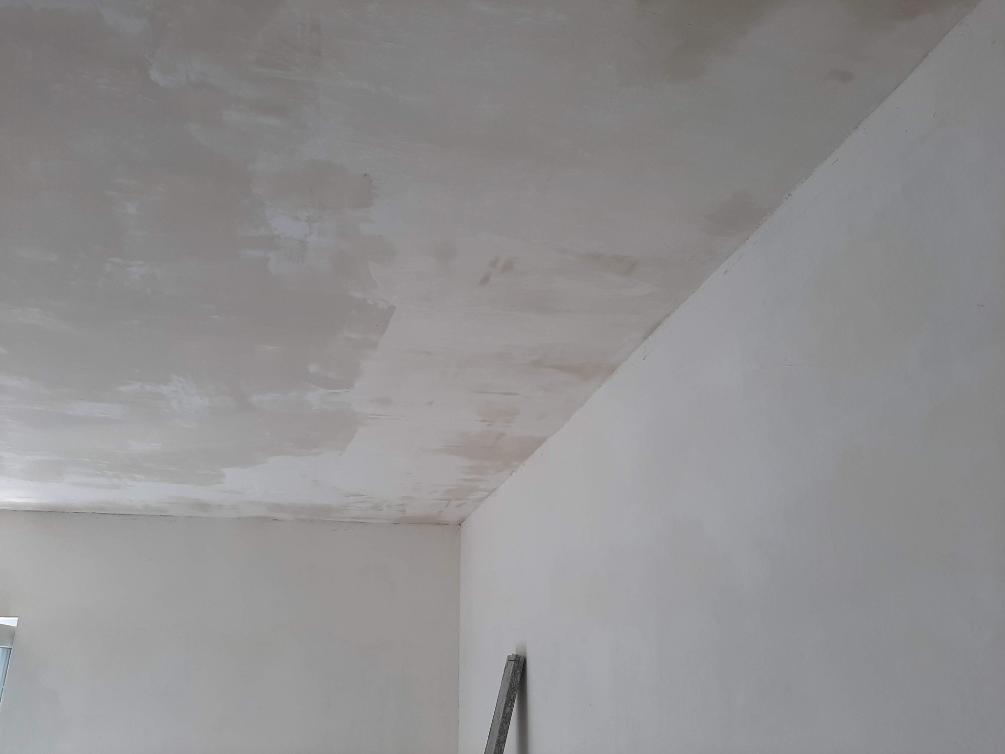 Ściana i sufit w trakcie prac wykończeniowych, widoczne ślady po nałożonej gładzi, szpachli, narożnik ściany i fragment drabiny malarskiej w dolnym rogu.
