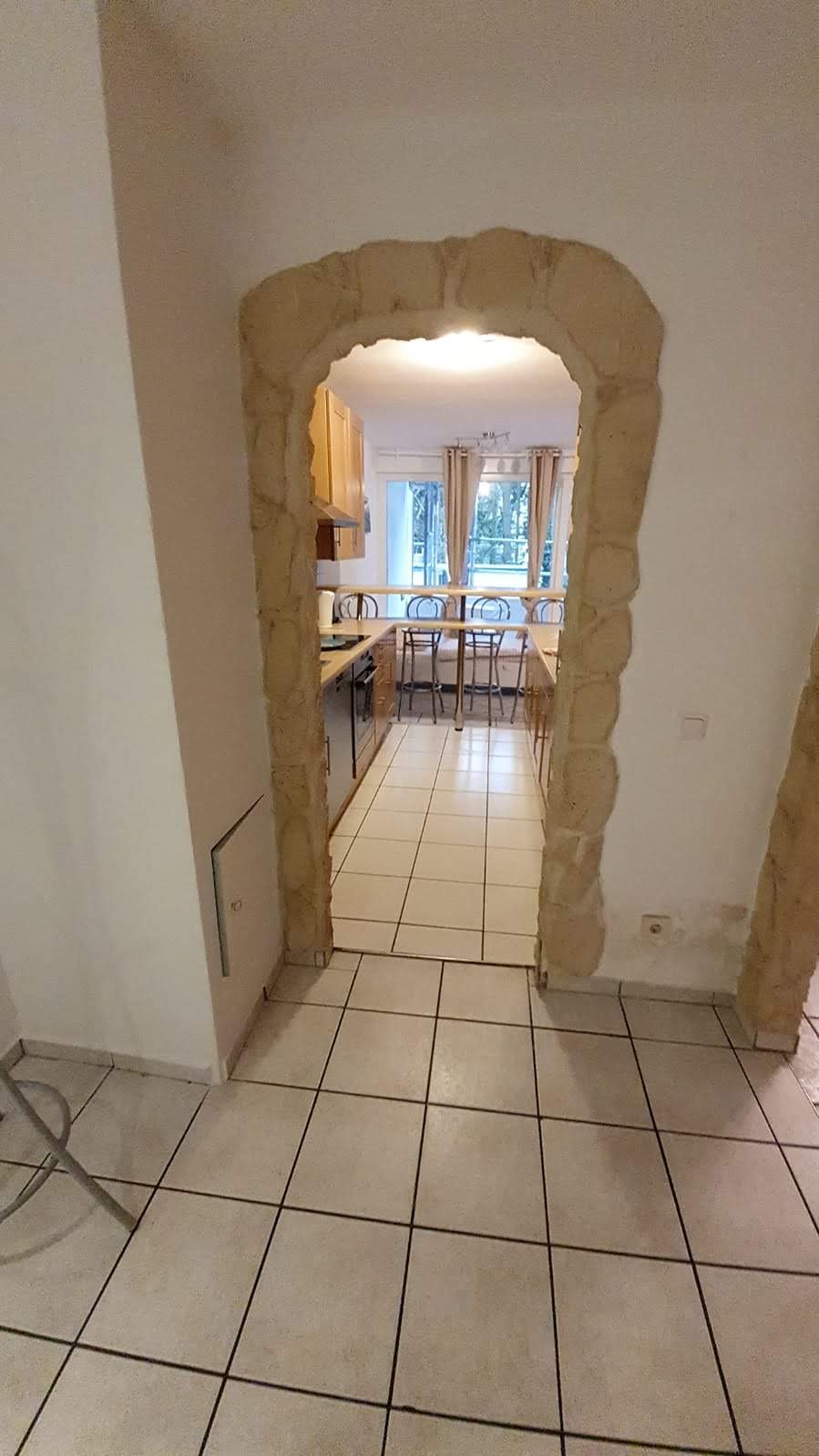 Widok na kuchnię przez kamienny łuk w ścianie, jasne płytki podłogowe, drewniane szafki kuchenne i krzesła barowe przy blacie.