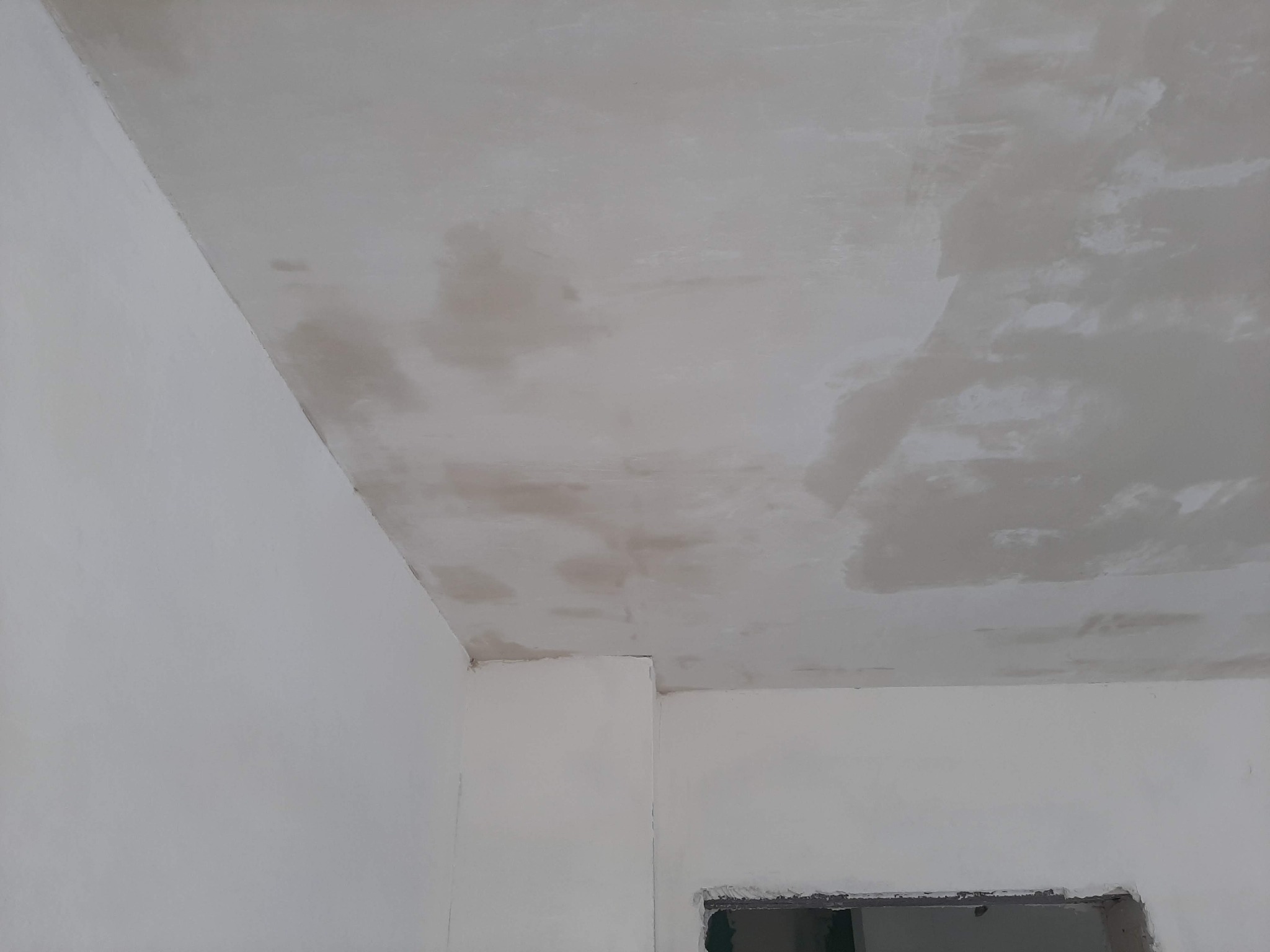 Nierównomiernie nałożona, szara gładź na suficie w trakcie schnięcia, widoczne fragmenty ścian w jasnym odcieniu szarości, przygotowane do dalszych prac wykończeniowych.