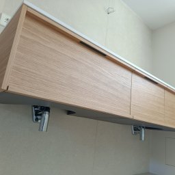 Glazur Usługi Budowlane Marcin Tokarz - Wisząca szafka łazienkowa z jasnego drewna z białym blatem, z czarnymi, podłużnymi uchwytami, zamontowana na tle jasnych płytek, widoczne chromowane syfony.