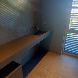 Glazur Usługi Budowlane Marcin Tokarz - Nowoczesna łazienka z długim, wiszącym blatem umywalkowym w kolorze antracytowym, szare płytki na ścianie z widoczną armaturą, marmurowa podłoga i okno z żaluzjami.