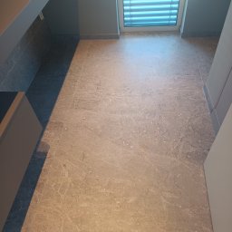 Glazur Usługi Budowlane Marcin Tokarz - Wykończona podłoga z płytek imitujących marmur w łazience, widok na okno z żaluzjami i fragment szafki pod umywalkę.