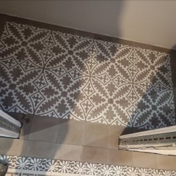 Glazur Usługi Budowlane Marcin Tokarz - Ułożona podłoga z płytek w stylu retro, z wzorem geometrycznym w odcieniach szarości i bieli, widoczne fragmenty listew przypodłogowych i ściany.