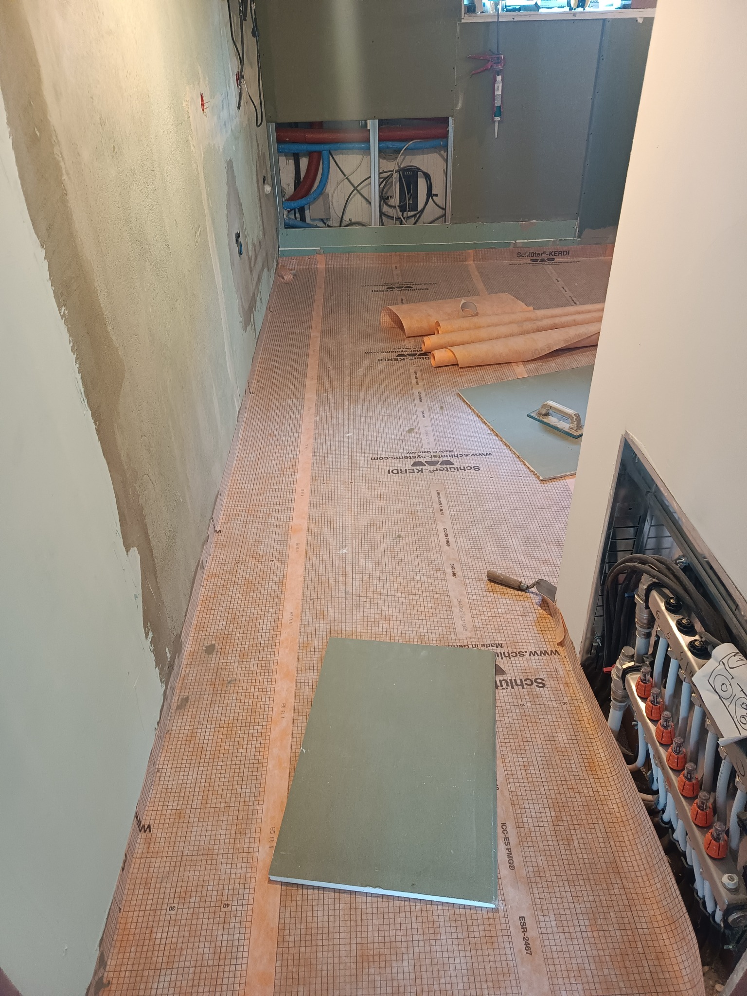 Przygotowanie podłoża pod płytki w łazience: rozłożona mata uszczelniająca Schluter Kerdi na podłodze, widoczne rury instalacji wodnej w ścianie i rozdzielacz ogrzewania podłogowego, płyta gipsowa...