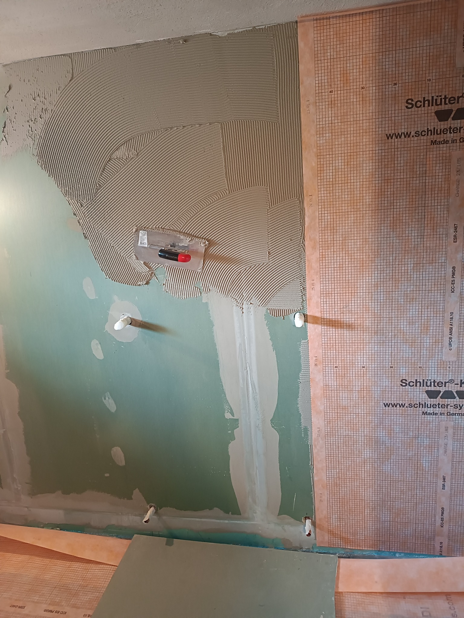 Ściana w trakcie przygotowania do układania płytek w łazience, z nałożoną warstwą kleju i przytwierdzoną membraną uszczelniającą Schlüter, widoczne wystające rurki instalacji wodnej i narzędzie...