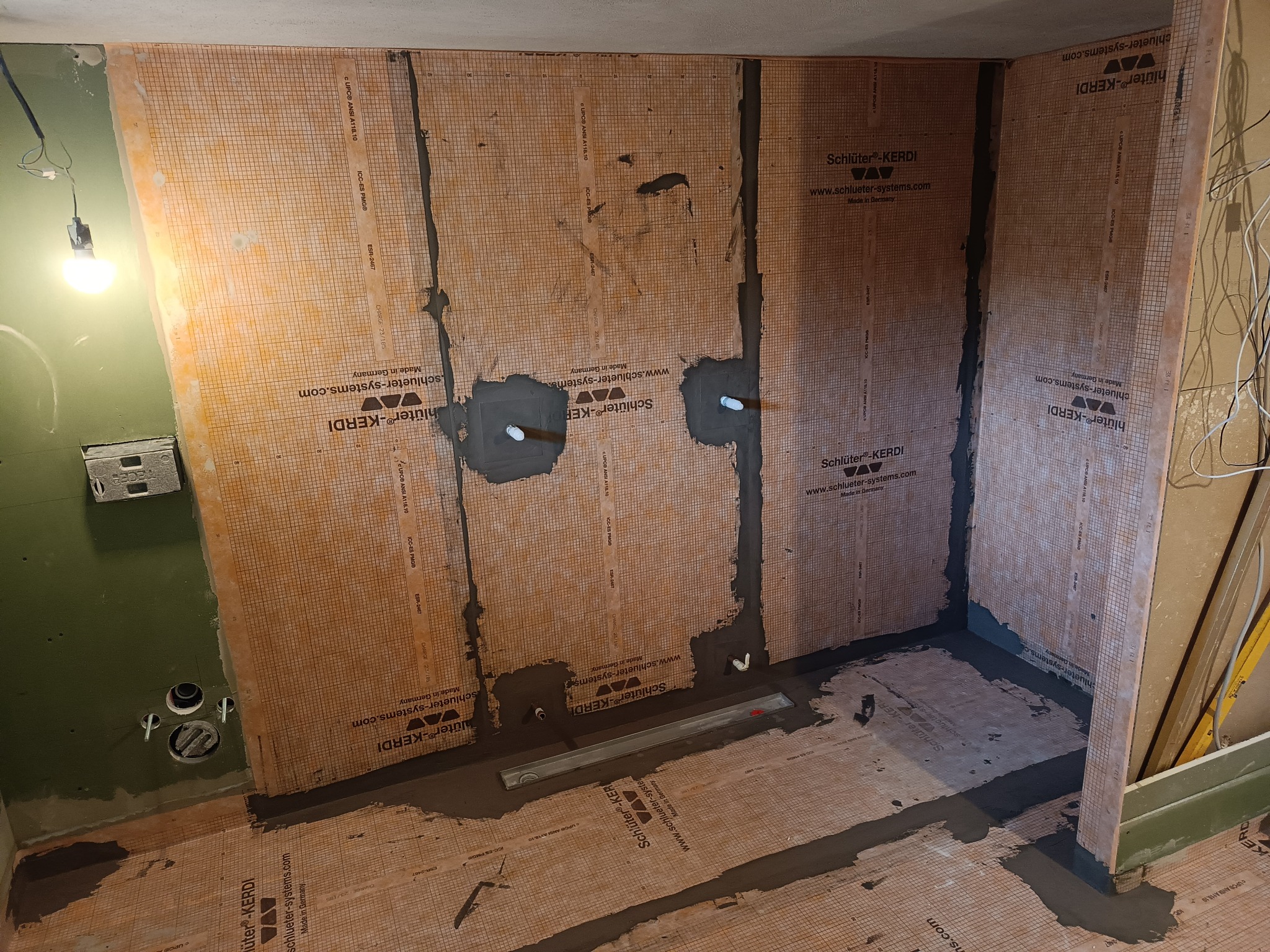Przygotowanie do hydroizolacji w łazience: widoczne membrany uszczelniające Schlüter-KERDI na ścianach i podłodze, z wklejonymi rurami i odpływem liniowym w podłodze, przed położeniem płytek.