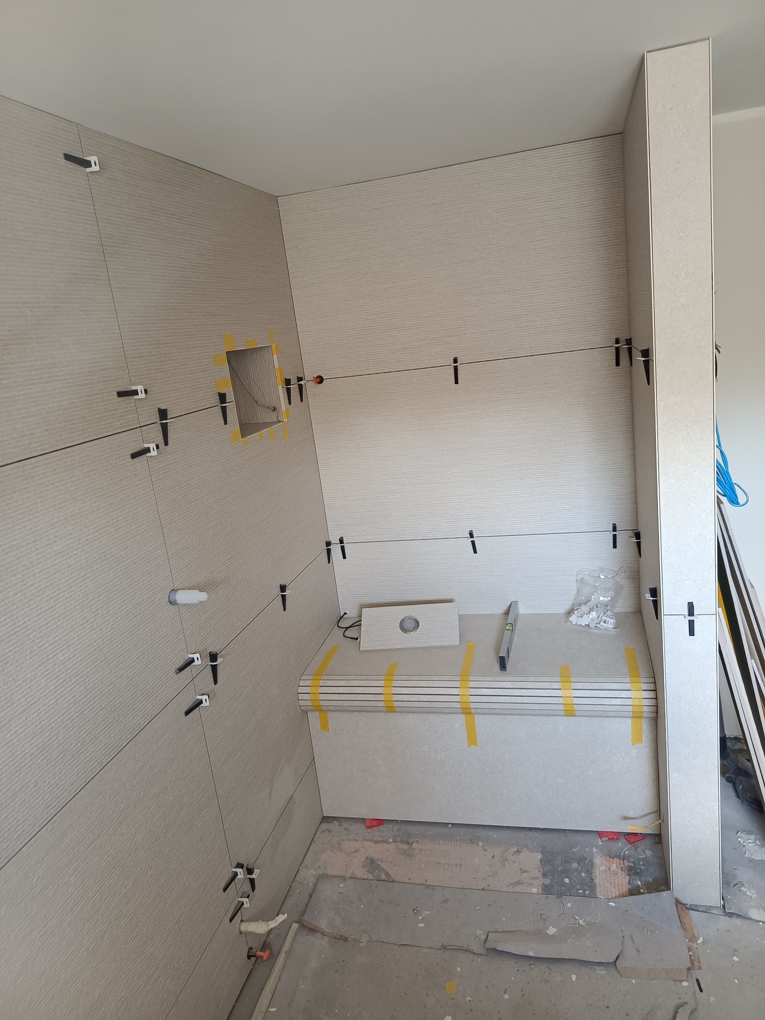 Montaż płytek ściennych z poziomymi paskami w łazience, widoczne krzyżyki dystansowe, poziomica oraz wycięty otwór na instalację.