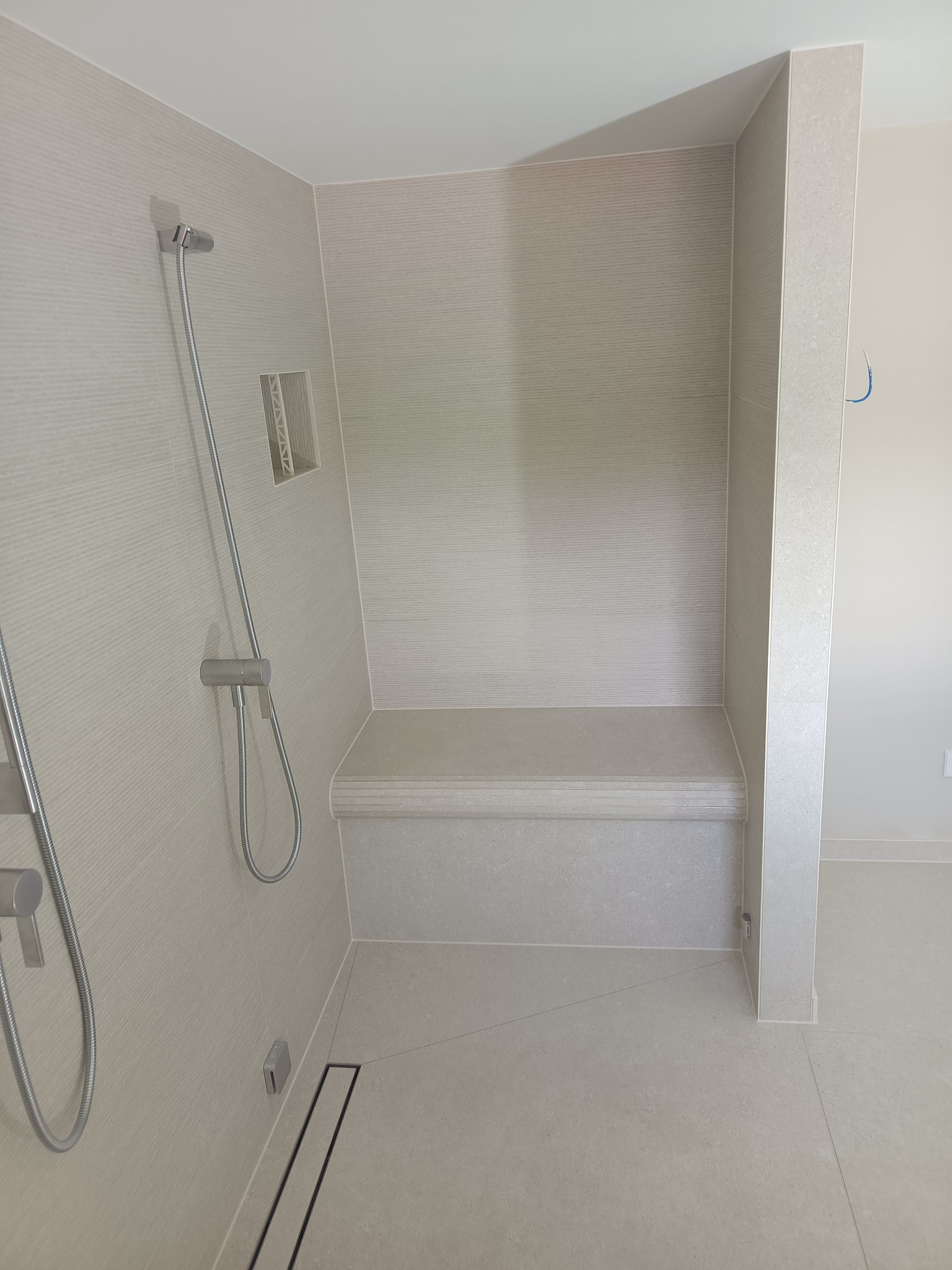 Nowoczesny prysznic z siedziskiem wyłożony beżowymi płytkami z delikatną fakturą, minimalistyczna armatura i odpływ liniowy.