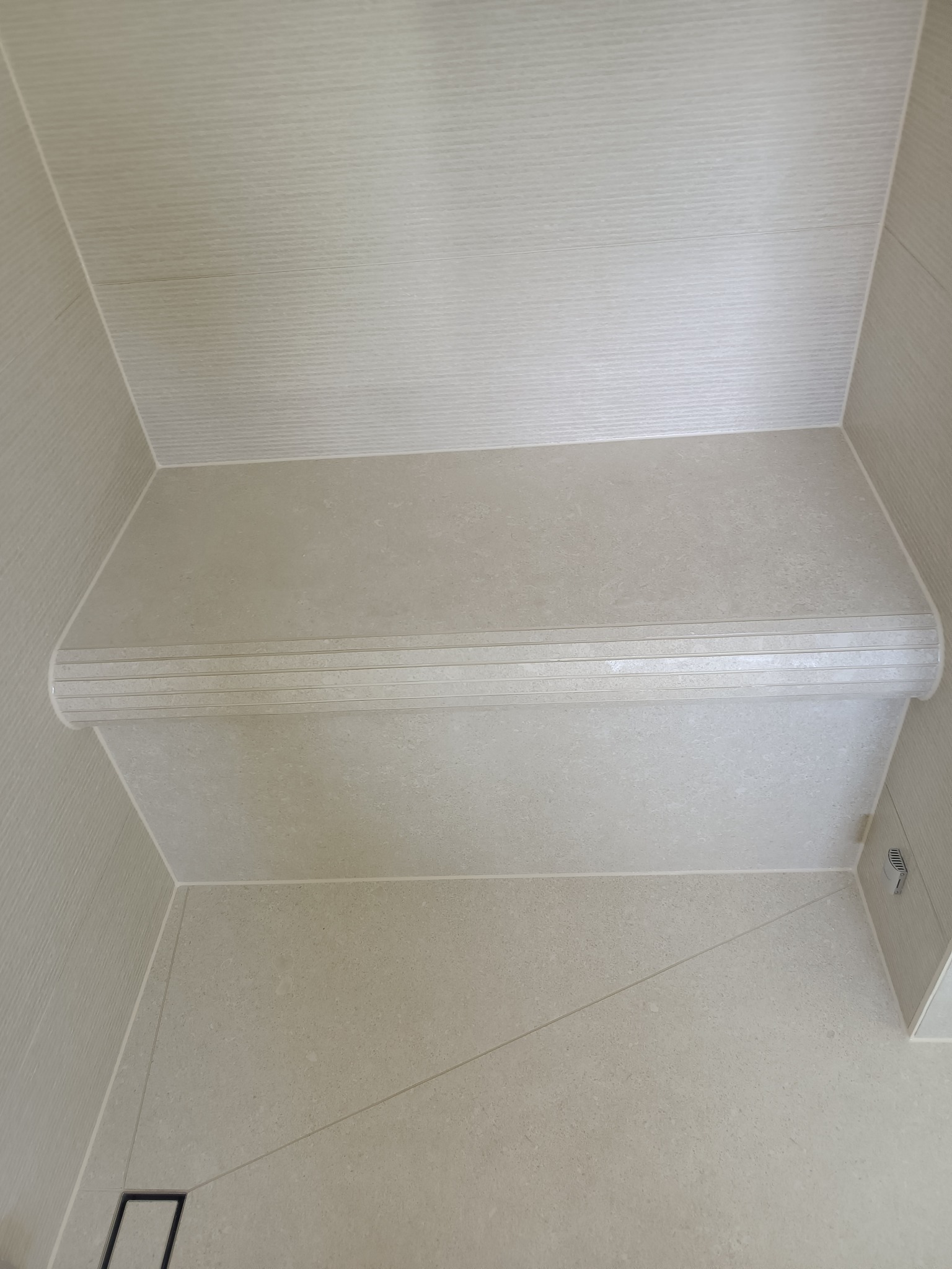 Wykończona wnęka prysznicowa z siedziskiem, wyłożona jasnymi płytkami imitującymi piaskowiec, z odpływem liniowym w podłodze.