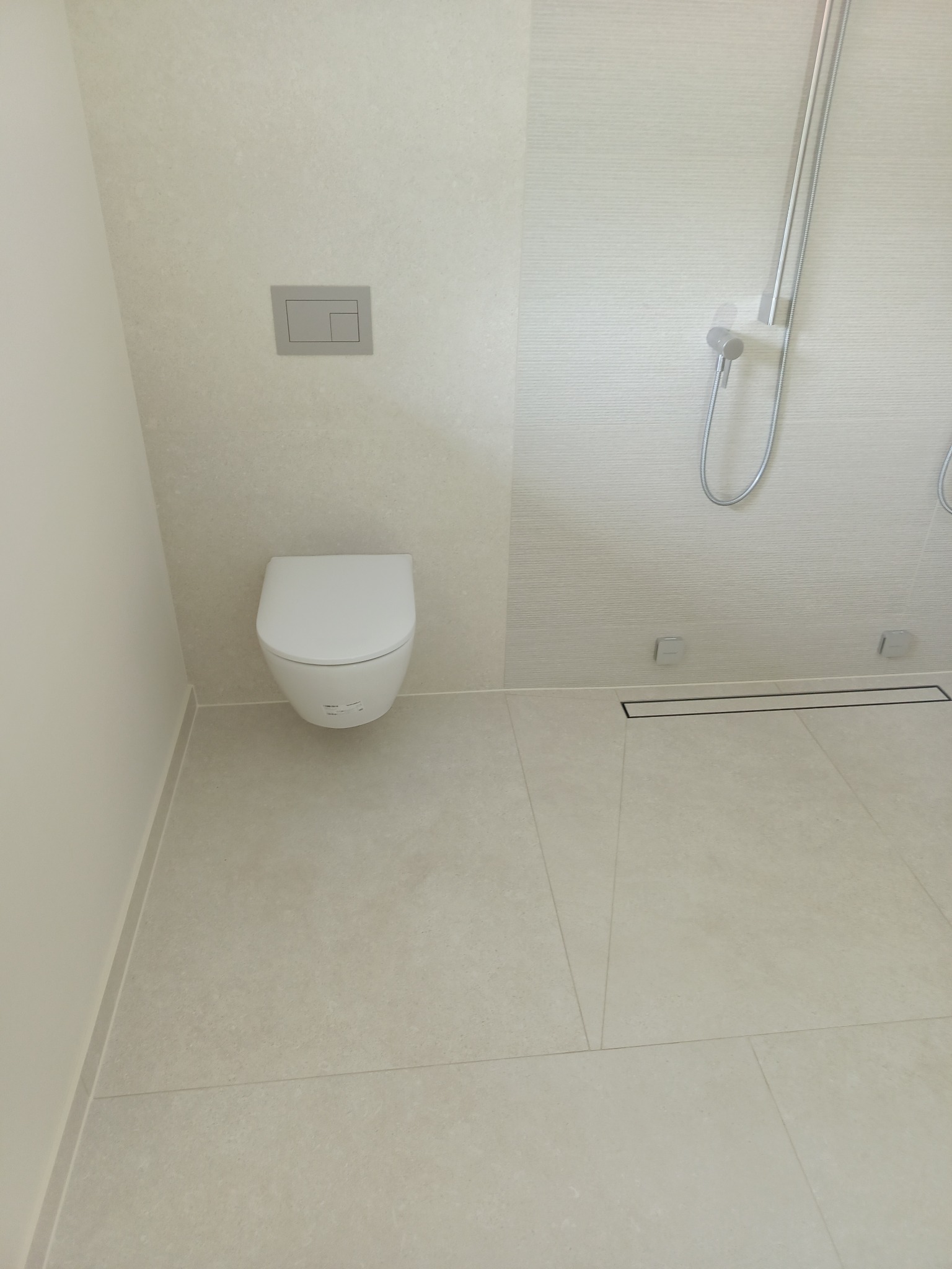 Nowoczesna łazienka z podwieszaną toaletą, minimalistyczną armaturą prysznicową i odpływem liniowym w podłodze wyłożonej jasnymi, kwadratowymi płytkami.