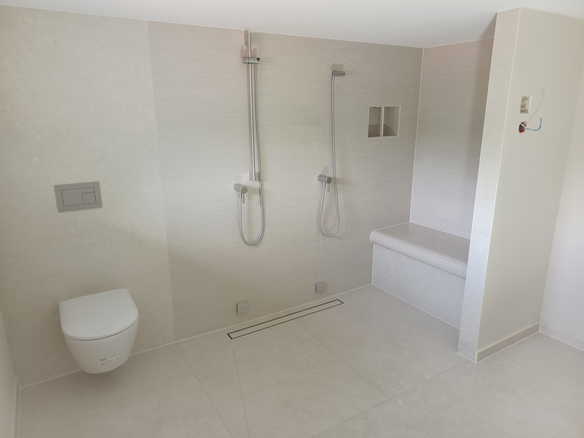 Jasna łazienka z dwoma natryskami, odpływem liniowym, podwieszaną toaletą i wnęką z siedziskiem, wykończona płytkami imitującymi tkaninę.