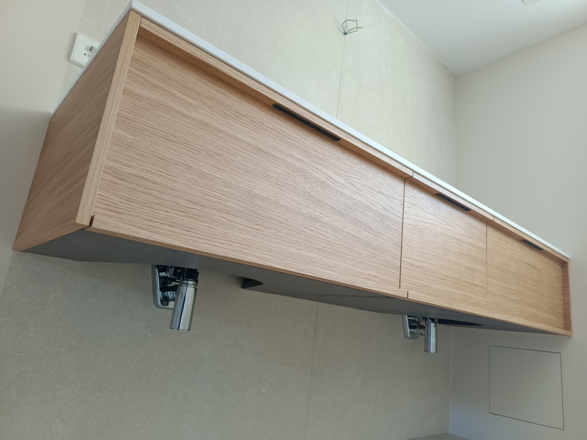 Wisząca szafka łazienkowa z jasnego drewna z białym blatem, z czarnymi, podłużnymi uchwytami, zamontowana na tle jasnych płytek, widoczne chromowane syfony.