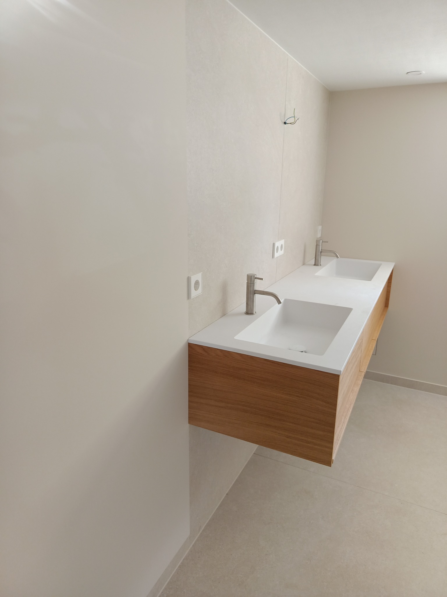 Nowoczesna łazienka z dwoma umywalkami nablatowymi na drewnianej szafce wiszącej, jasne płytki na ścianie, widoczne gniazdka elektryczne i wystające przewody.