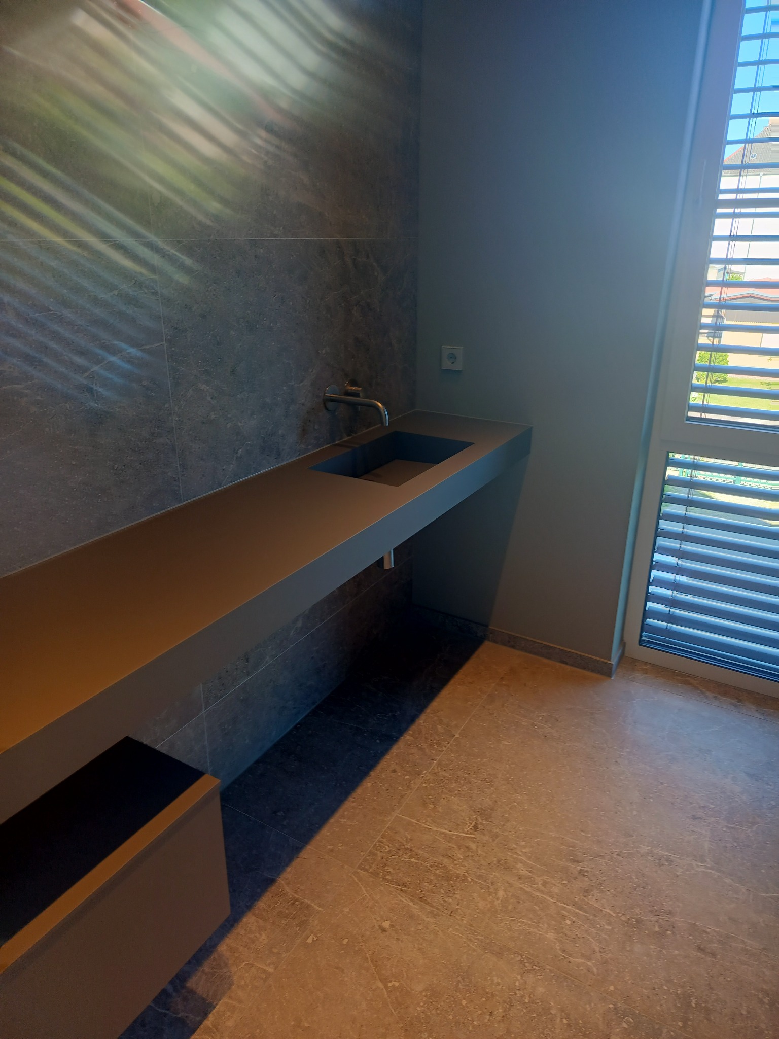 Nowoczesna łazienka z długim, wiszącym blatem umywalkowym w kolorze antracytowym, szare płytki na ścianie z widoczną armaturą, marmurowa podłoga i okno z żaluzjami.