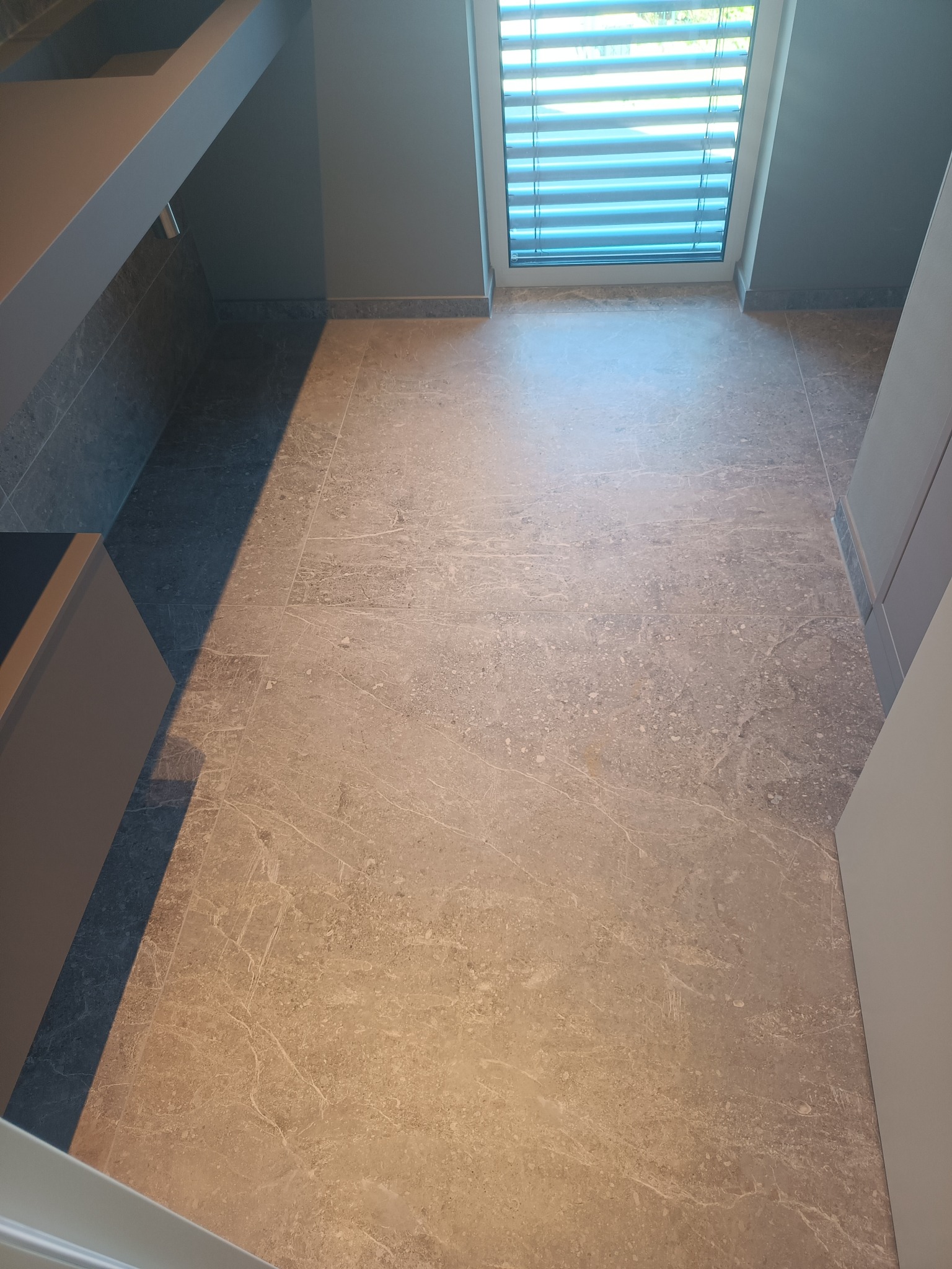 Wykończona podłoga z płytek imitujących marmur w łazience, widok na okno z żaluzjami i fragment szafki pod umywalkę.