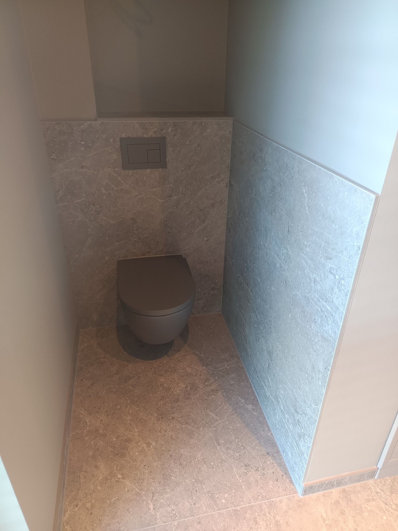 Nowoczesna toaleta w zabudowie, szara miska WC i marmurowe płytki na ścianach oraz podłodze, minimalistyczny przycisk spłuczki.