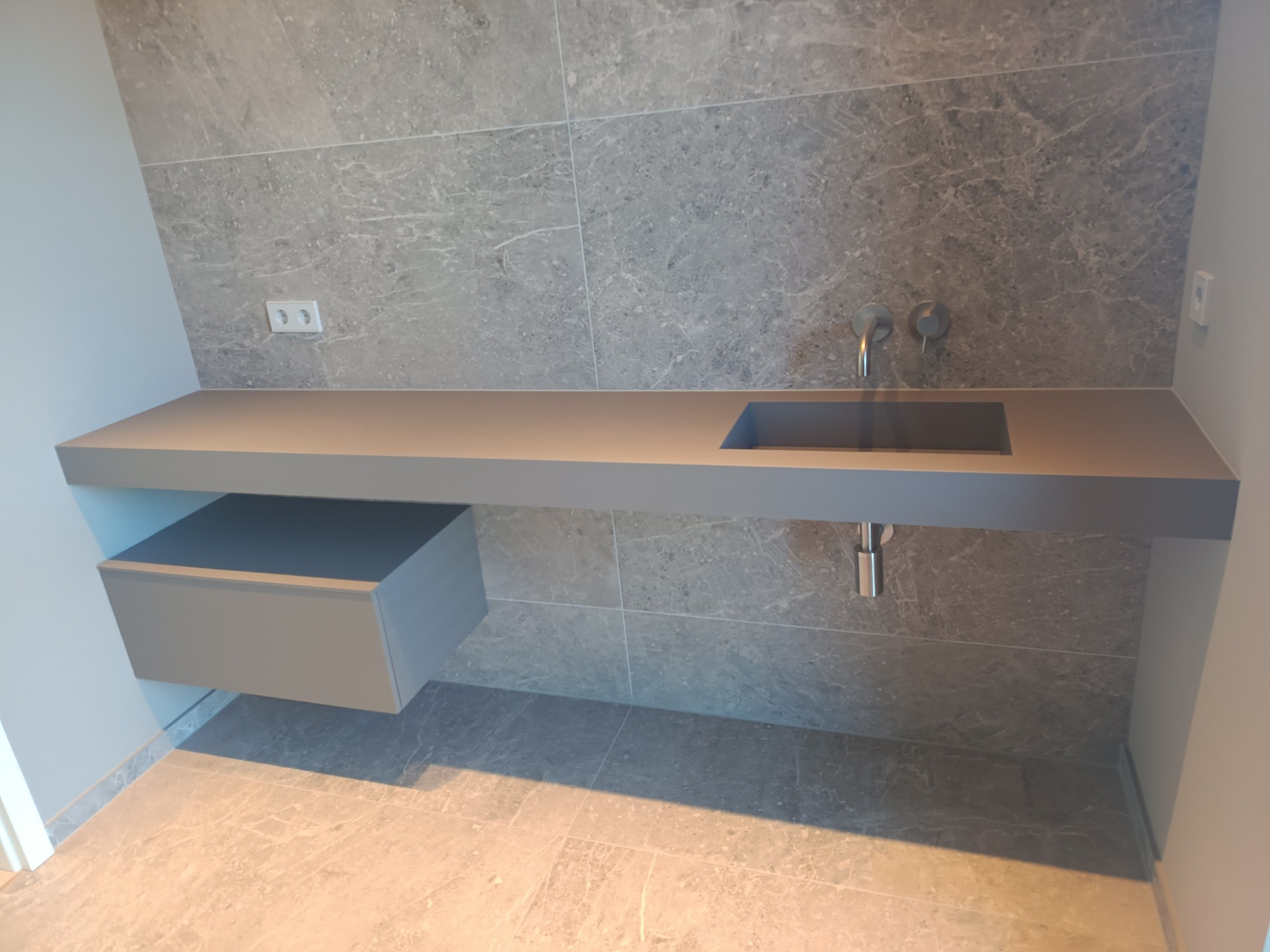 Szara łazienka z umywalką wpuszczoną w blat, bateria ścienna, szara płytka imitująca marmur na ścianie i podłodze, szafka pod umywalką z jedną szufladą.