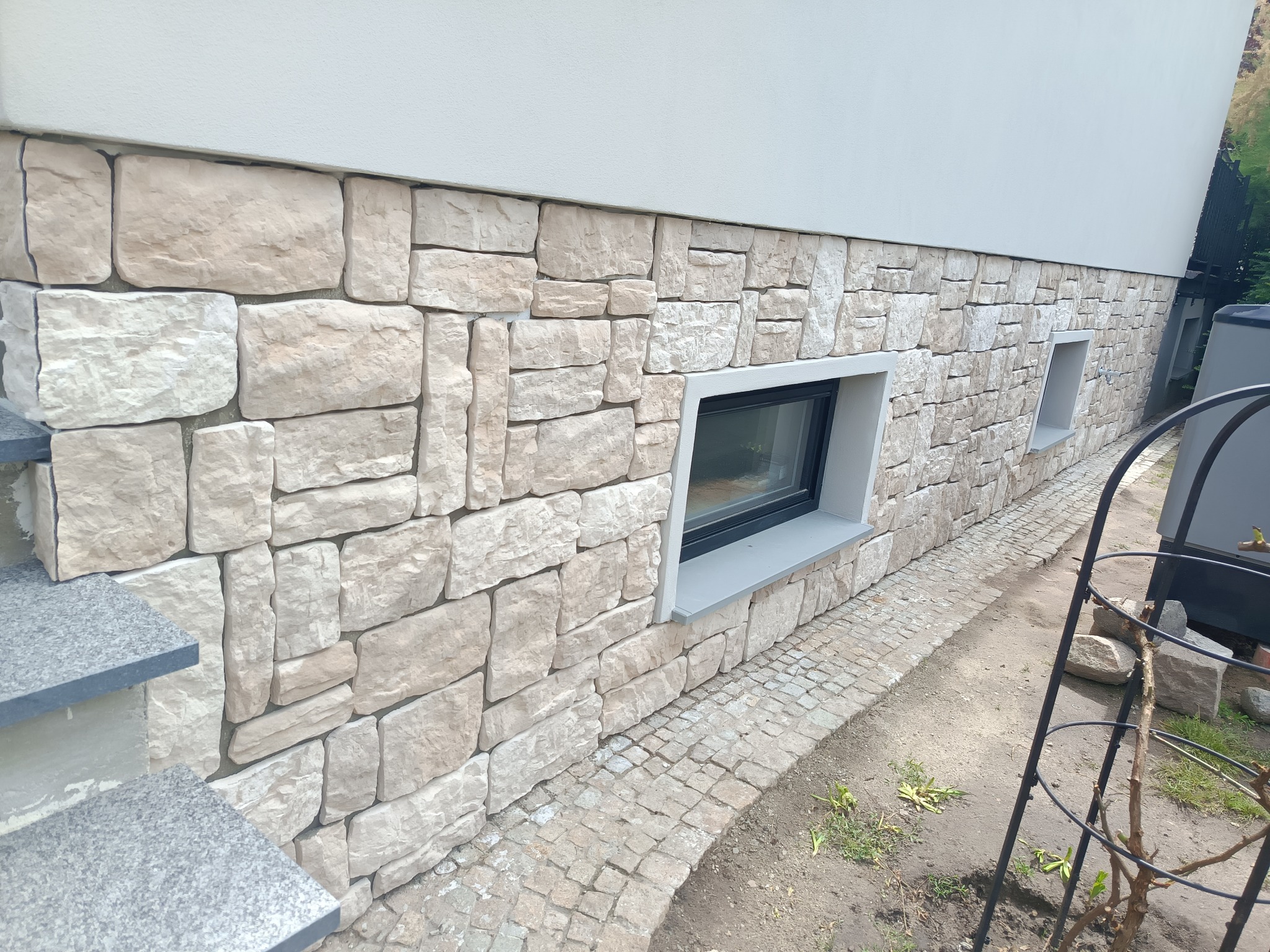 Elewacja budynku wykończona kamieniem naturalnym w odcieniach beżu i szarości, z widocznymi dwoma oknami piwnicznymi i fragmentem schodów wejściowych.