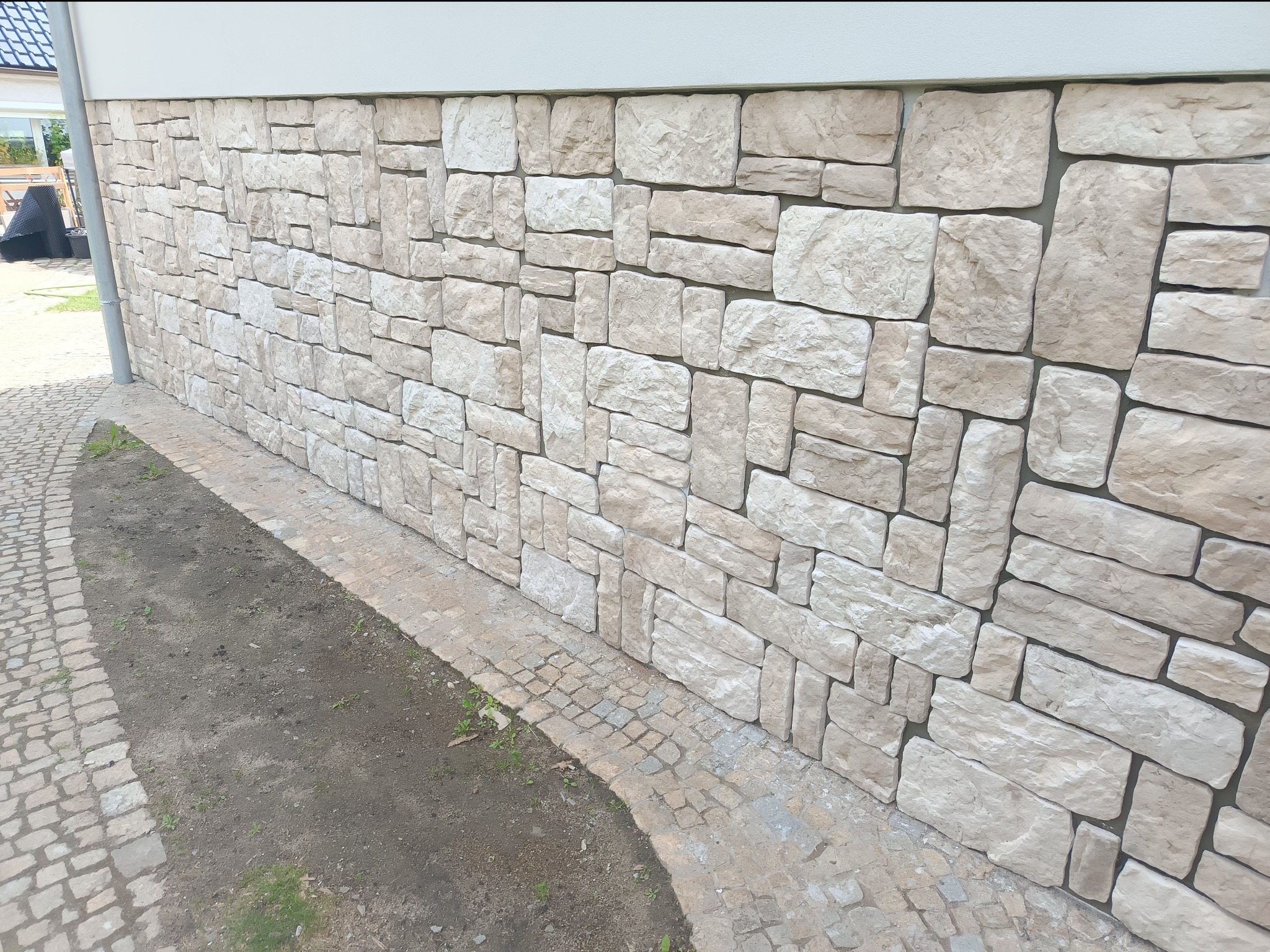 Ściana budynku wykończona dekoracyjnym kamieniem elewacyjnym w odcieniach beżu i szarości, z widocznym fragmentem chodnika z kostki brukowej i rabatą ziemi wzdłuż ściany.