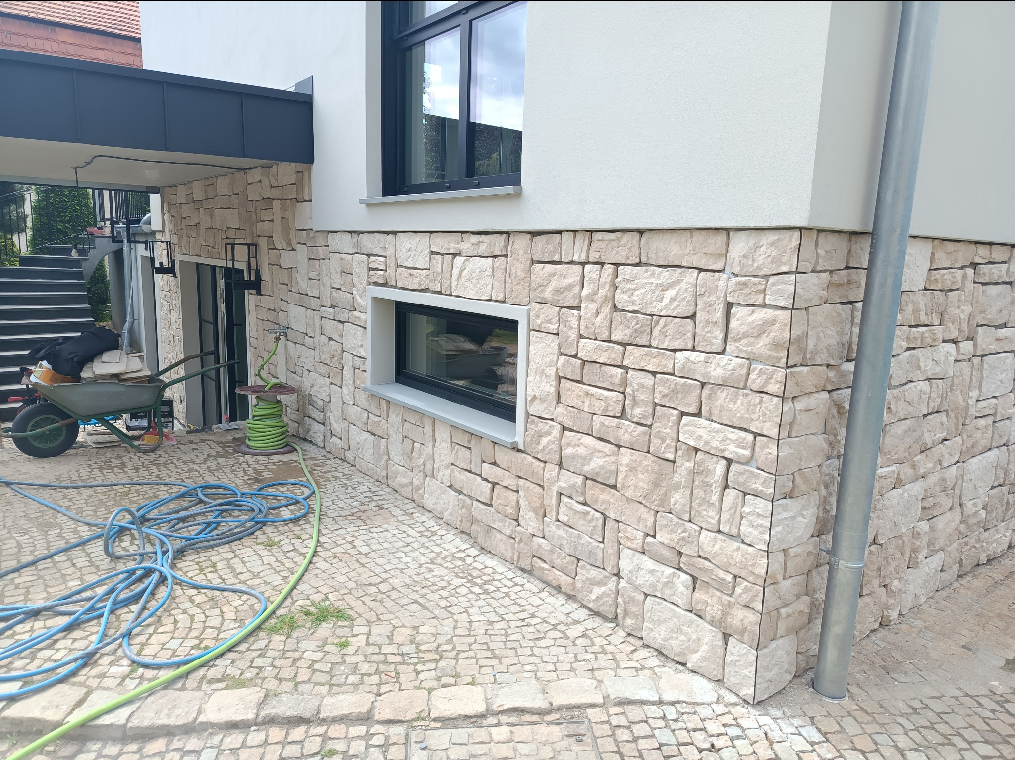 Elewacja domu wykończona jasnym kamieniem naturalnym, widoczny fragment ściany z oknem, obok leży zwinięty niebieski wąż ogrodowy, a w tle taczka i zielony wąż na szpuli.