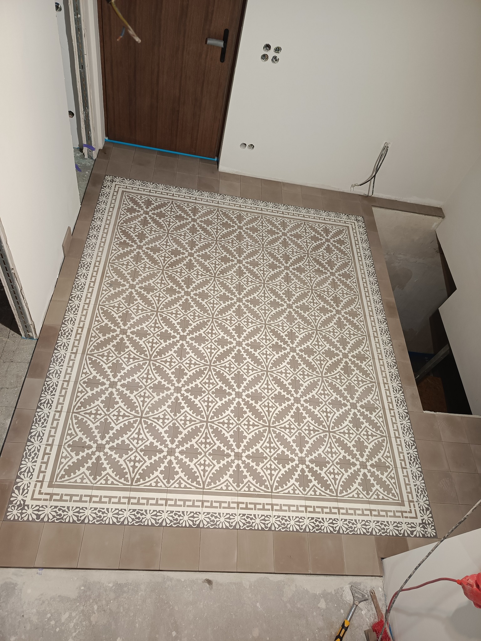 Wzorzysta podłoga z płytek w odcieniach beżu i bieli, ułożona w nowym pomieszczeniu, widoczne niedokończone ściany i instalacje elektryczne w suficie.