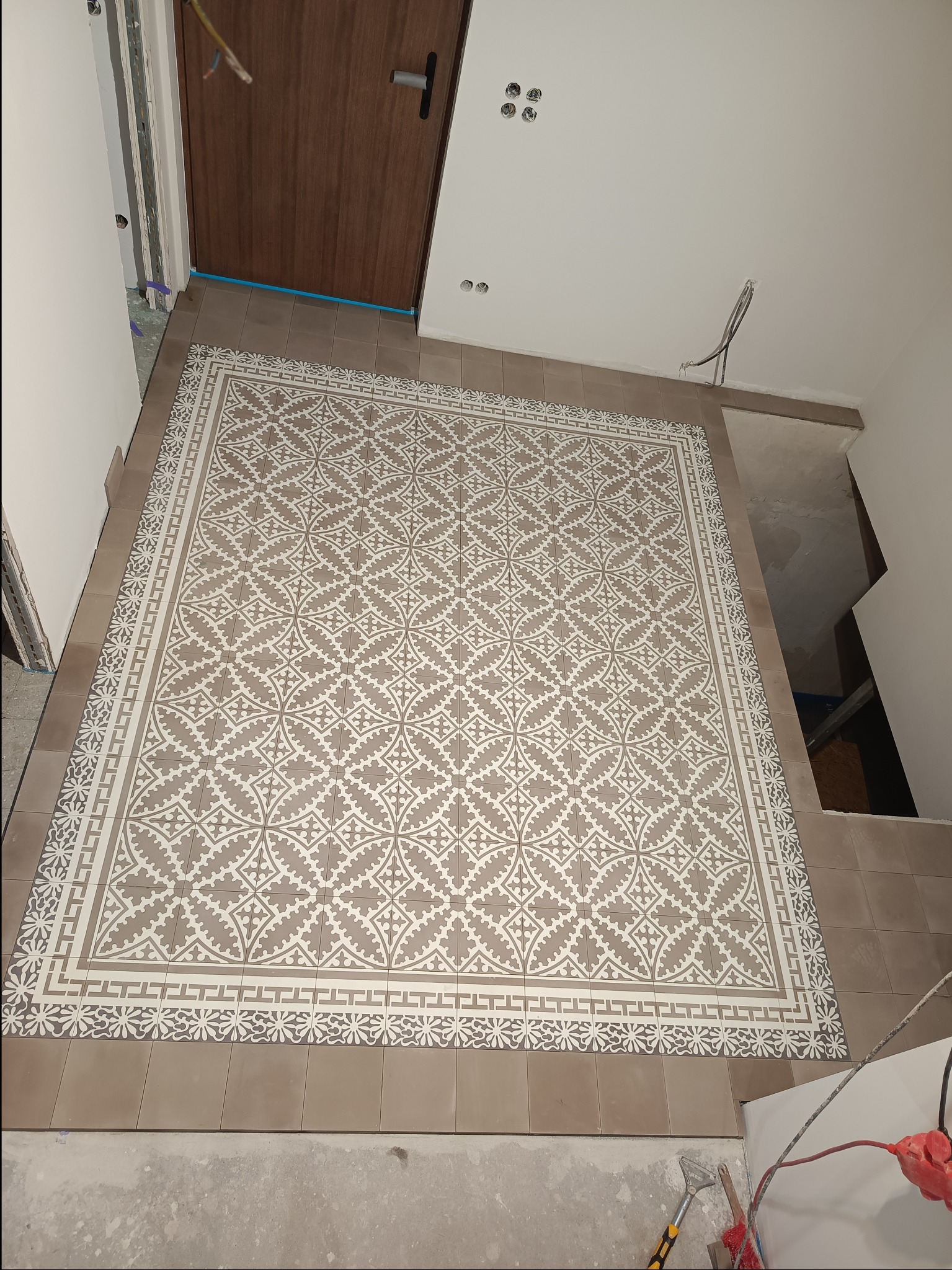 Ułożona podłoga z płytek o wzorze geometrycznym w odcieniach beżu i bieli, widoczne narzędzia i kable elektryczne w tle, fragment drzwi wejściowych i surowy stan wykończenia ścian.