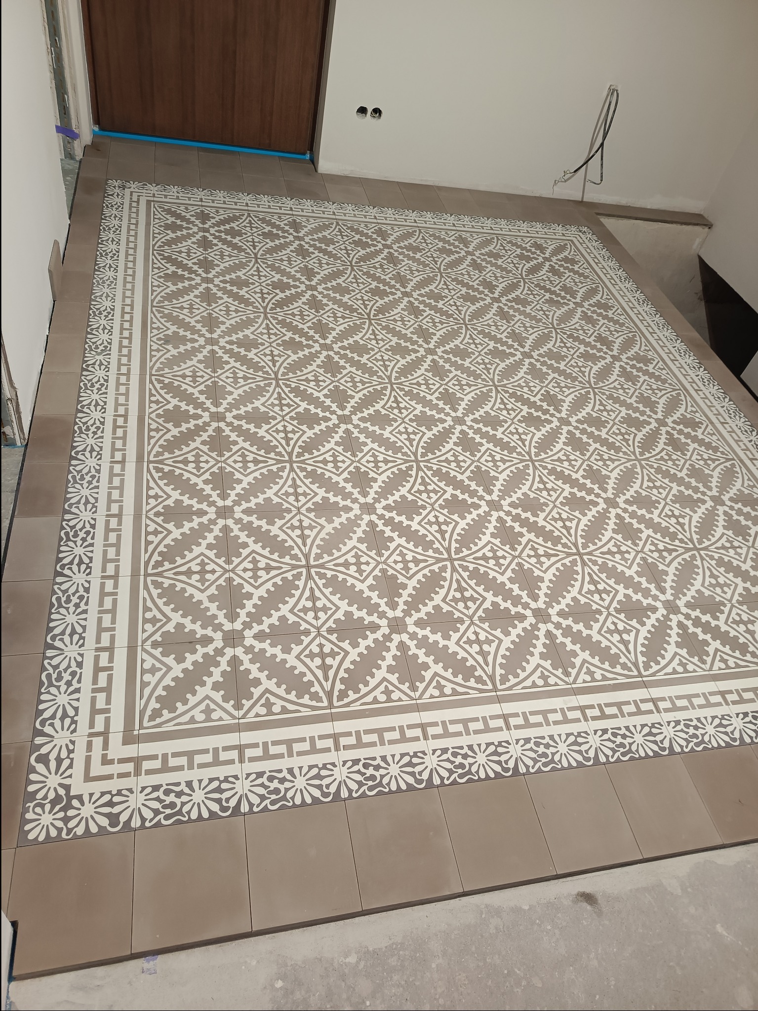 Ułożona podłoga z płytek ceramicznych w geometryczne wzory w odcieniach beżu i bieli, widoczne niedokończone krawędzie i wystające kable, wskazujące na trwający proces wykończeniowy.