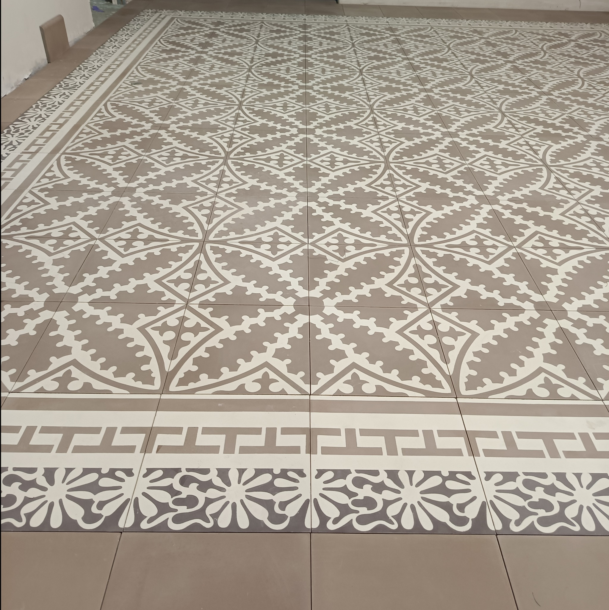 Ułożona podłoga z płytek w odcieniach beżu i bieli, z dekoracyjnym wzorem geometrycznym i motywem kwiatowym na obrzeżach.