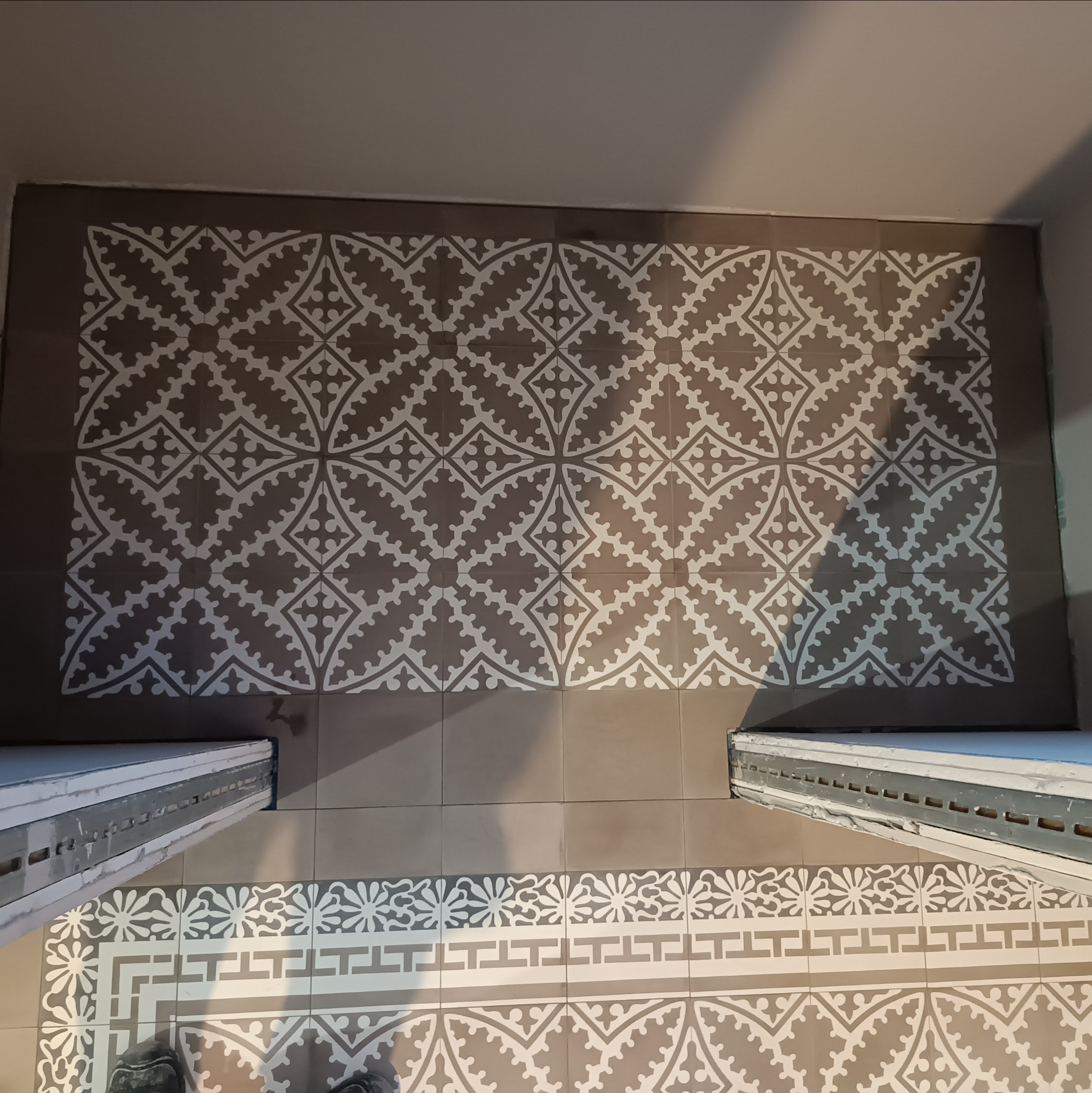 Ułożona podłoga z płytek ceramicznych w łazience, wzór geometryczny w odcieniach szarości i bieli, widoczne fragmenty progów drzwiowych i butów