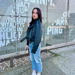 Kobieta w czarnej skórzanej kurtce i dżinsach stoi na chodniku przed szklaną ścianą z napisem.