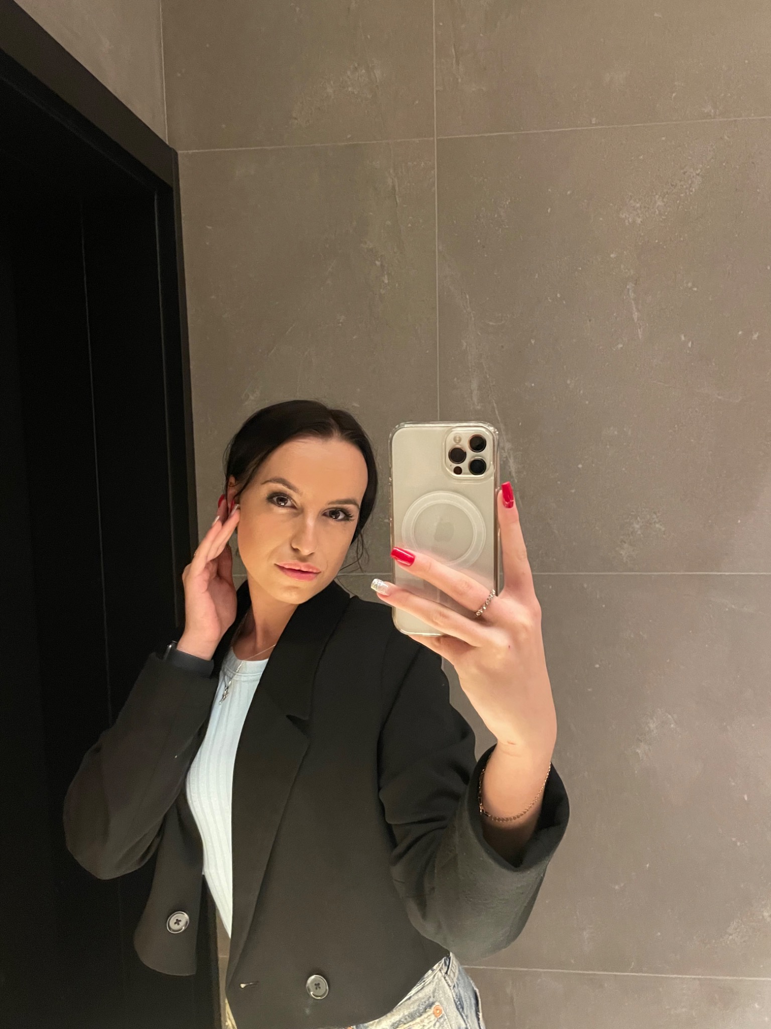 Kobieta w czarnej marynarce robi selfie telefonem z przezroczystym etui na tle szarej ściany.