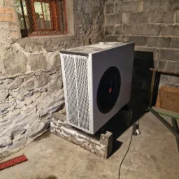 Biała pompa ciepła zamontowana na betonowych wspornikach w piwnicy o kamiennych ścianach i małym oknie z kratą.