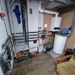 Piwnica z kotłem gazowym, zasobnikiem wody, rurami grzewczymi i narzędziami na podłodze. Widoczny także fotel i środki czystości.