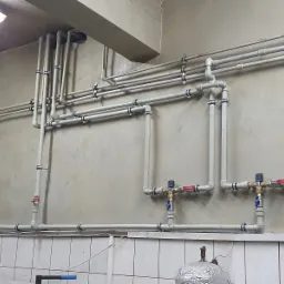 Szare rury instalacyjne z zaworami i mocowaniami na ścianie z szorstkim tynkiem i białymi płytkami.