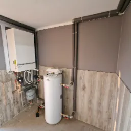 Nowoczesna kotłownia z piecem gazowym i zasobnikiem ciepłej wody, rury w izolacji termicznej prowadzone wzdłuż ściany wykończonej panelami imitującymi drewno, narzędzie akumulatorowe leżące...