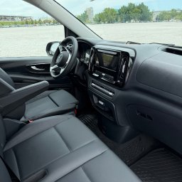 MBVBUS - Wnętrze busa Mercedes Vito: czarna skórzana tapicerka, deska rozdzielcza z ekranem dotykowym, widok na parking i budynki w tle przez szybę. Detal.