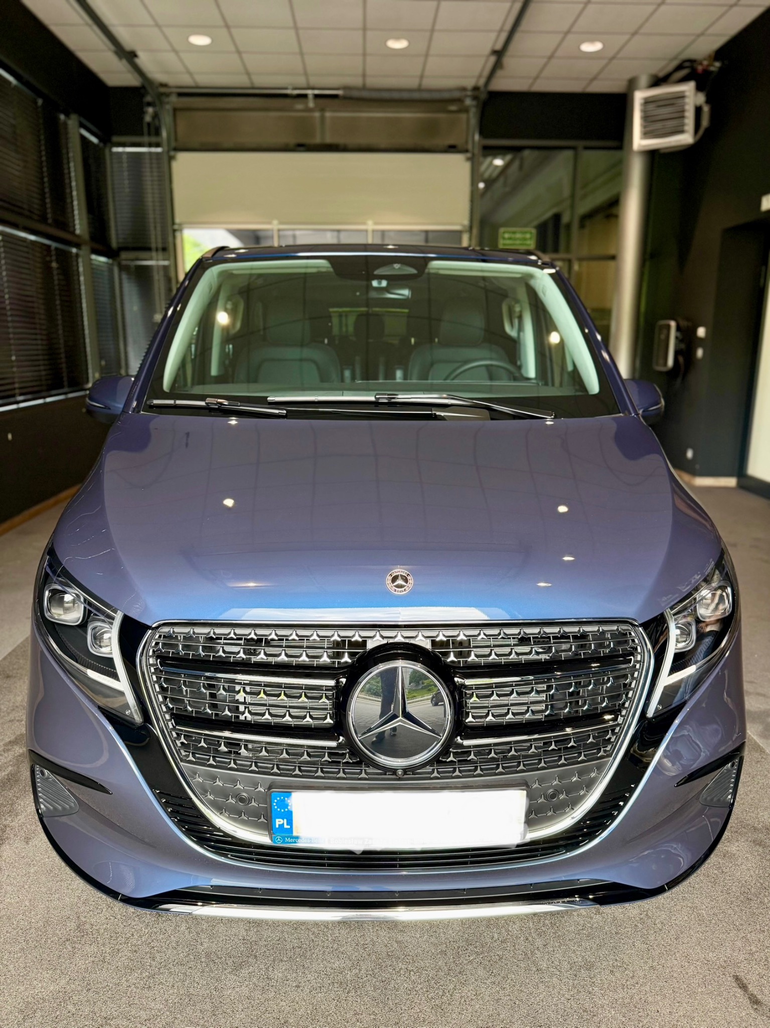 Niebieski Mercedes-Benz EQV z chromowanym grillem i reflektorami LED, zaparkowany w salonie samochodowym. Widoczny fragment tablicy rejestracyjnej z oznaczeniem PL.