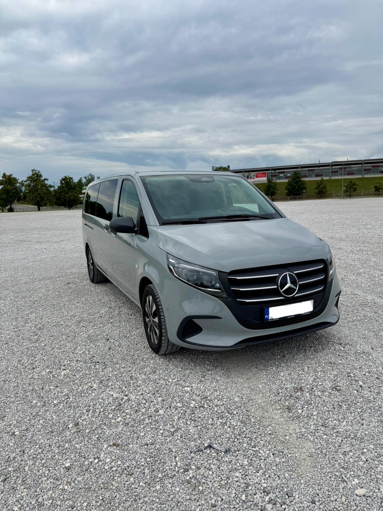 Szary Mercedes Vito na żwirowym parkingu, pochmurne niebo w tle. Auto z przodu, tablica rejestracyjna zasłonięta.