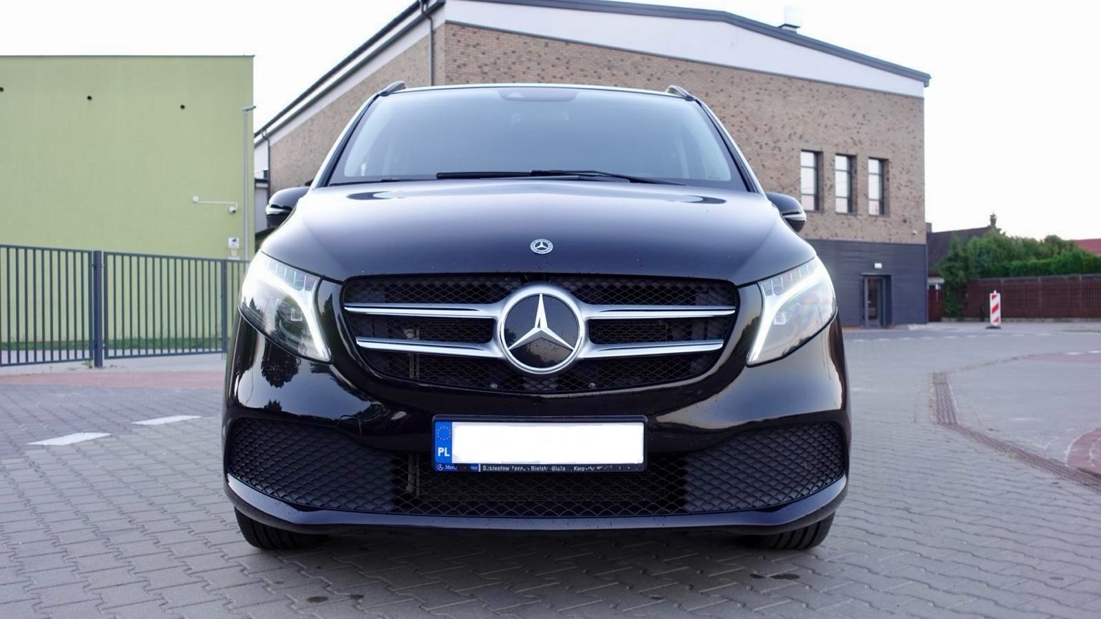 Czarny Mercedes V-klasa z przodu, z widoczną atrapą chłodnicy i reflektorami, zaparkowany na kostce brukowej, budynek w tle. Tablica rejestracyjna zasłonięta.